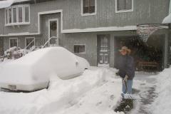 Kathy shoveling snow from the driveway, next to a car that's buried in snow