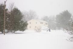 view across the street shows little sign of plowing; neighbor using snowblower; snow falling