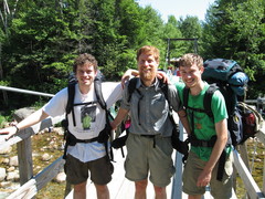 three guys with backpacks smiling, on a suspension bridge
